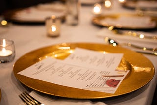 A gold dinner plate on a formal dining table with a menu in the foreground is accented by candlelight and out-of-focus dinnerware.