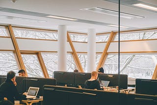 An open place office setting, with multiple workers at each of several bench style desks.