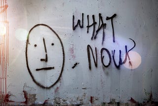 Photo of a peeling gray concrete wall. There is graffiti of a face with a neutral expression and the words “What Now?” beside it.