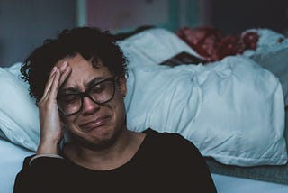 A middle-aged woman sits on the floor beside her bed with her hand to her forehead, crying.