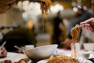 a table packed with plates and bowls of chinese food with two hands, each holding a pair of chopsticks from there are noodles hanging.