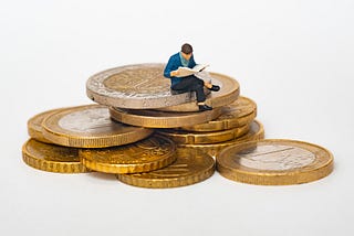 A toy figure of a man seated on a stack of coins and reading a book