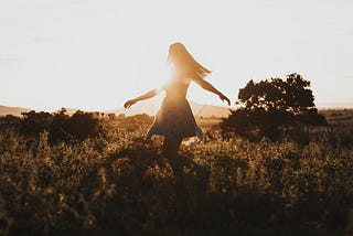 A woman dances in a field, the sun low in the sky behind her.