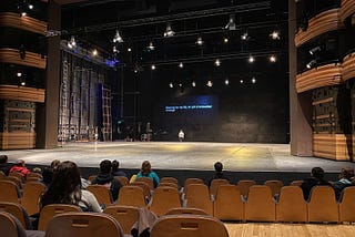 A big stage with not much in it apart from one person standing towards the back of it and a screen reading “Dancing for my life: An act of embodied courage”. Some people sat on brown seats in the audience. House lights are on.