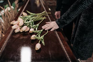 A woman’s hand lays gently on the lid of a polished casket. It’s dark brown in colour, and pale pink tulips are left by the mourners on the lid.