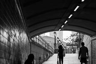 A black and white photo of a homeless person sitting under a bridge with two people walking by.