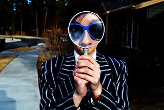 A person who is wearing black suit and black spectacle cornered with blue colour, holding a magnifier lens from both hands.