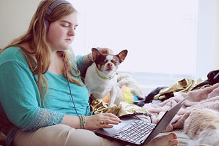A woman in a blue shirt sits on a bed with a laptop on her lap. She pets a small dog with her left hand and appears sad or frustrated.