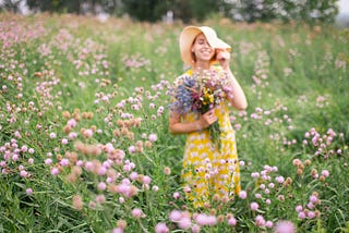 A woman smiles while holding a bouquet in a green field with purple wild flowers.