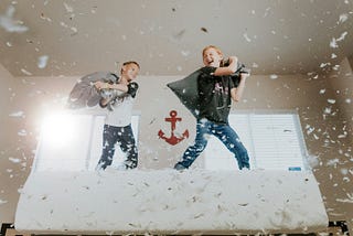 Tow boys pillow-fighting on a bed with feathers flying.