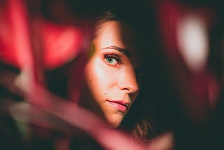 A woman peeks out from some blurry red foliage, one piece of which is heart-shaped