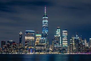 The Manhattan skyline is shown, lit up against the dark night sky, the water in the forefront.