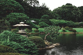 Jardim aberto, com árvores médias e uma lamparina japonesa em volta de um pequeno lago, em um dia nublado.