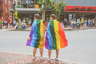 two people on a sidewalk, wrapped in rainbow flags