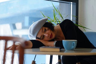 Photograph of a young woman in a beret waiting in a coffee shop, resting her head on her arm.