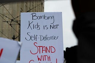 Written in black and red ink, a white sign at a protest reads: Bombing Kids is NOT Self-Defense STAND WITH GAZA!