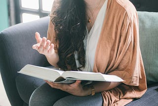 torso of long-haired woman in white t-shirt and beige cardigan sitting on couch with an open book