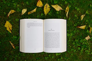 A book that is open lays on the green grass with yellow leaves above it.