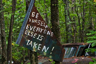 A picture of a rustic black poster in a forest, it has white writings reading “Be yourself everyone else is taken.” Used to depict the idea of being authentically you.