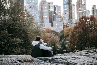 A couple is sitting on a large rock looking at a skyline with trees in front.
