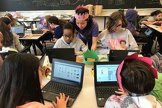 Classroom of students working on Scratch using personal computers. A teacher in pink crown assists students in the upper left side of the image.