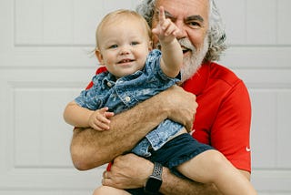 A bearded gray haired man wearing a red shirt holds a baby in front of a garage.