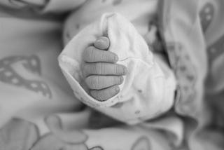 Black and white photo with closeup of an infant’s hand in an oversized sleeve. The baby’s sleeve with hand is surrounded by a blanket with non discernable pattern in gray tones.