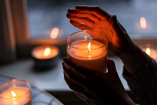 Image of a burning candle in a glass holder, held by a hand and the other hand shading the flame. Other burning candles in holders fill the space around the center image.