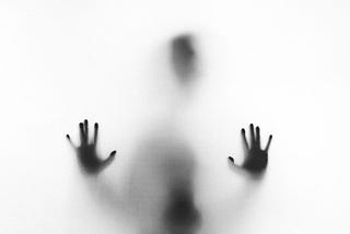 A figure through frosted glass, only a vague shape of a body and head, and two hands against the glass.