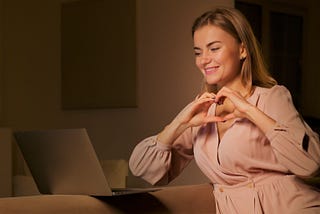 A woman making a heart shape with her hands in front of a laptop