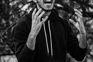 A young man passionately expresses anger or frustration, gesturing dramatically against a blurred natural background