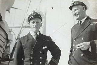 Phot depicts two naval captains standing outside in the sun, in their uniforms, laughing and one is smoking.