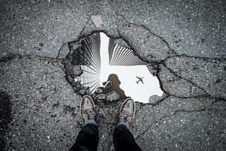 A person gazing into a puddle of water in cracked cement, the puddle reflecting an airplane and skyscrapers.