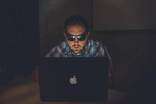 Image of a person sitting behind a laptop in the dark with sunglasses on