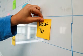 A person places a sticky note on the wall with the words ‘HOW TO’, signaling a focus on instructions, guidance, or problem-solving.