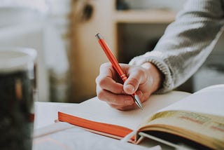 An orange pen held by a hand about to write on grid-lined paper.