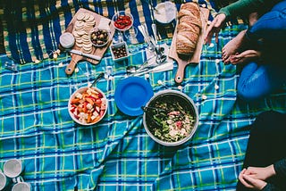 picnic on a blue and white checkered blanket, with bread, salad, fruit, olives, crackers, and cracker sides