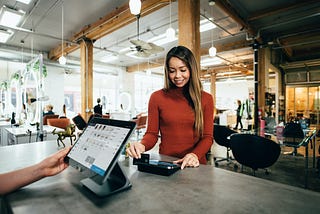 A woman with long highlighted hair and a burnt orange long-sleeved knit top swipes her debit or credit card at a payment terminal opposite a person across the counter standing in front of a tablet point-of-sale device in an open cafe or lounge. you can only see the other person’s forearm and hand holding one corner of the terminal.