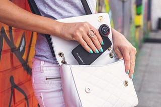 A white handbag is slung across a woman’s body. It is open and the woman is slipping her phone into it. She has turquoise nail polish on and behind her is a wall with red and green graffiti.