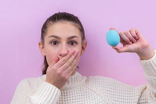 Surprised brunette girl stands on a pink background and holds an Easter egg. Easter. Easter bunny. Easter egg. Holiday. Advertising. Place for an inscription. Postcard. Brunette girl. Smirk