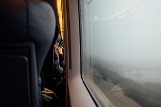 A person sitting inside a train gazing outside the window at the foggy fields.