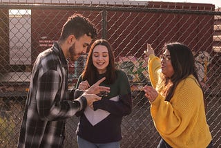 Three young people stand in front of a chainlink fence. Left is a young man with a beard, wearing a plaid shirt and holding a smartphone. Middle is a young woman with long brown hair, wearing a dark shirt, and right is another young woman with long dark hair, wearing glasses and a mustard yellow sweatshirt. They are all looking at the smartphone and smiling.