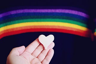 A person holding a pink heart with a cloth rainbow in the background.