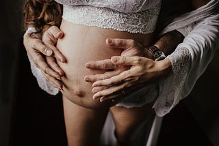 The doctor is in, and he’s bred me. Pregnant erotica. Doctor breeding patient erotica. A man stand behind a woman with their hand on her pregnant belly. The pregnant woman is wearing white lace.