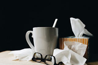 A box of tissues, some loose tissues, a mug, and a pair of glasses all clustered on a rattan mat.