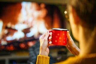 A woman drinking from a red cup in front of a fireplace. Cancelation of Stephen Colbert, The Late Show Stephen Colbert, Strike Force Five, Entertainment, Myers, Stewart, Late Night, Kimmel, talk show, comedy, first amendment, when is late show ending? Oliver, Fallon, democracy, mental health, coffee, Paramount, CBS, opinion, stress, mental decline, sucking taxpayers, Colbert last show.
