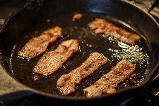 Depicted is a skillet with some bacon being fried. It is a perfect snack when on the keto diet due to it’s high fat content.