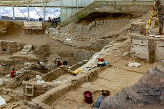 Photograph of an archaeological excavation in progress, showing exposed stretches of stone buildings and other features at different depths, with archaeologists digging or surveying separate portions of the site.