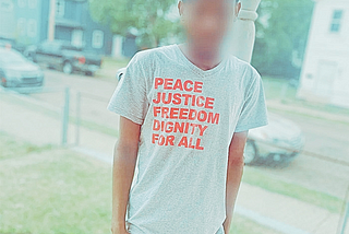 A young man with a blurred face wears a t-shirt that says “Peace, Justice, Freedom, Dignity for All.”