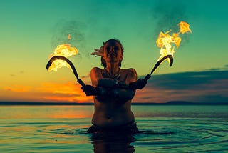 A person silhouetted against the sunrise, stands waist deep in the ocean holding two flaming torches with arms crossed.